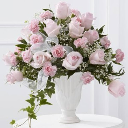 Elegant sympathy flowers with soft pink roses and carnations, complemented by baby&rsquo;s breath, ivy, and greenery, beautifully arranged in a white pedestal vase with a sheer ribbon for a comforting touch.