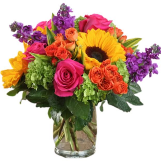 Colorful summer arrangement featuring sunflower, fuchsia roses, orange roses, purple stock, and greenery in a glass vase.