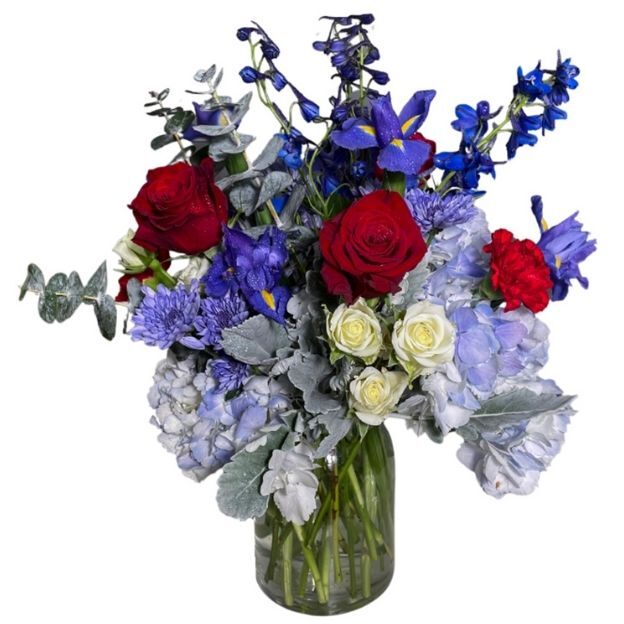 Elegant mix of red roses, blue delphinium and iris, blue hydrangea, white spray roses, and silver foliage in a clear vase.