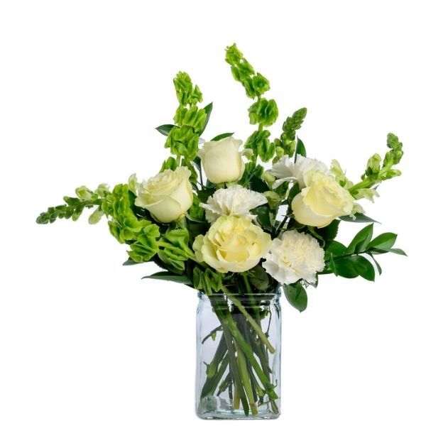 White roses and carnations with green bells of Ireland and foliage in a square glass vase.