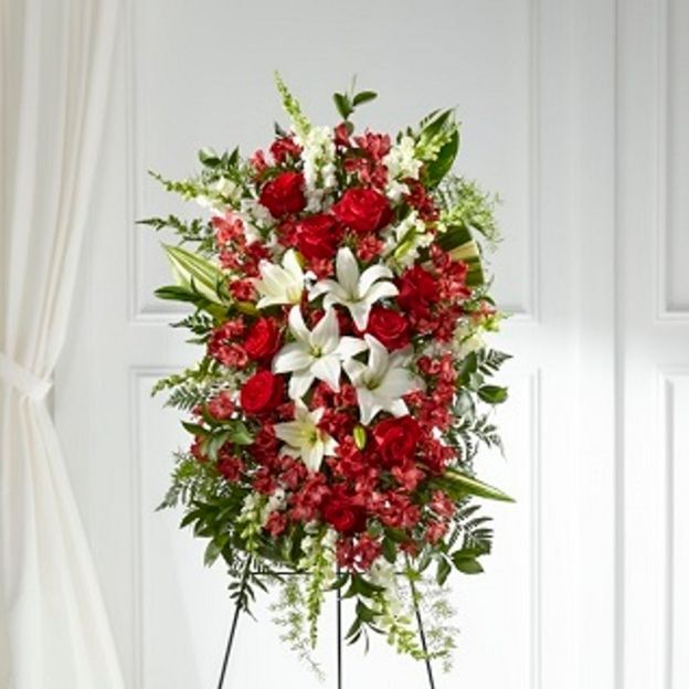 Red and white standing spray with roses, lilies, and greenery on an easel in a bright chapel.