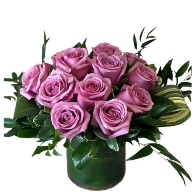A bouquet of pink-lavender roses arranged in a round glass vase with green leaves and decorative foliage.