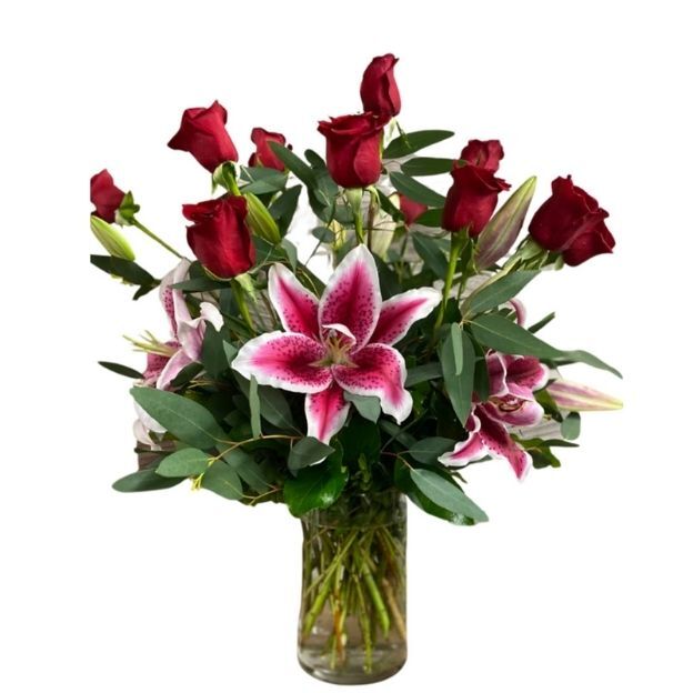 A bouquet of red roses and pink lilies arranged in a clear vase with lush greenery and decorative foliage.