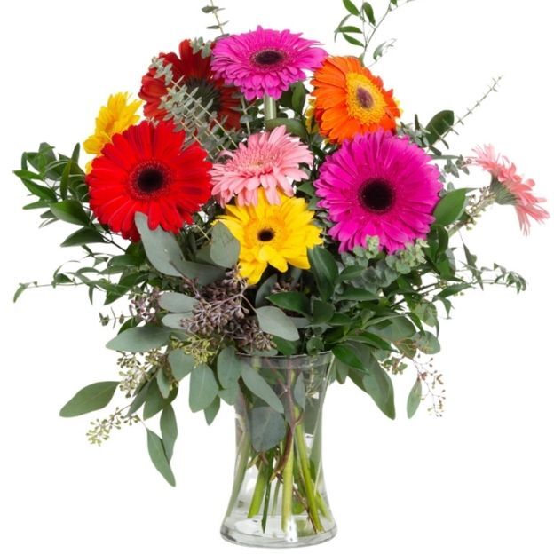 A bouquet of vibrant gerbera daisies in pink, red, orange, and yellow, arranged in a clear glass vase with lush green foliage.