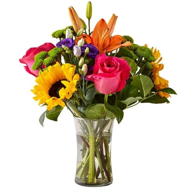 Mixed flower arrangement in a clear vase featuring pink roses, orange lilies, sunflowers, and purple blooms.