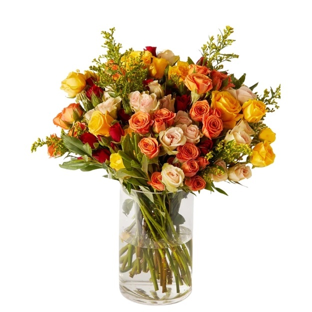 Full bouquet of yellow, orange, cream, and red roses with leafy greenery and yellow sprigs in a clear glass vase.