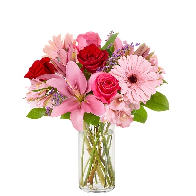 Pink and red bouquet featuring an open pink lily, pale pink daisies, and roses in a tall cylindrical glass vase.