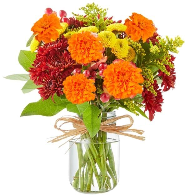 Bright fall bouquet of orange carnations, red chrysanthemums, yellow poms, and greens arranged in a clear jar vase.