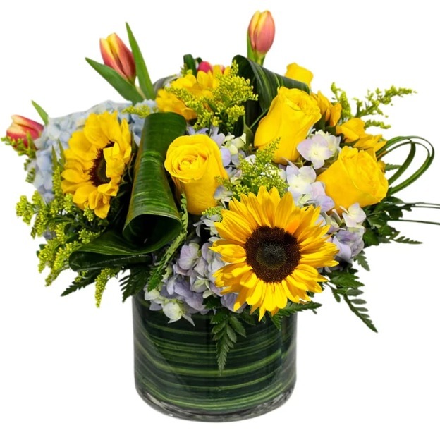 Yellow sunflowers and roses with peach tulips, pale blue hydrangeas, and curled leaves in a leaf-lined glass vase.