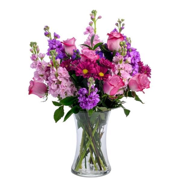 Pink roses with purple stock and asters, arranged with greenery in a flared clear vase.