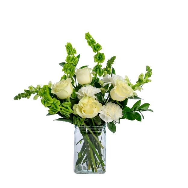 White roses and carnations with green bells of Ireland arranged in a clear square glass vase.