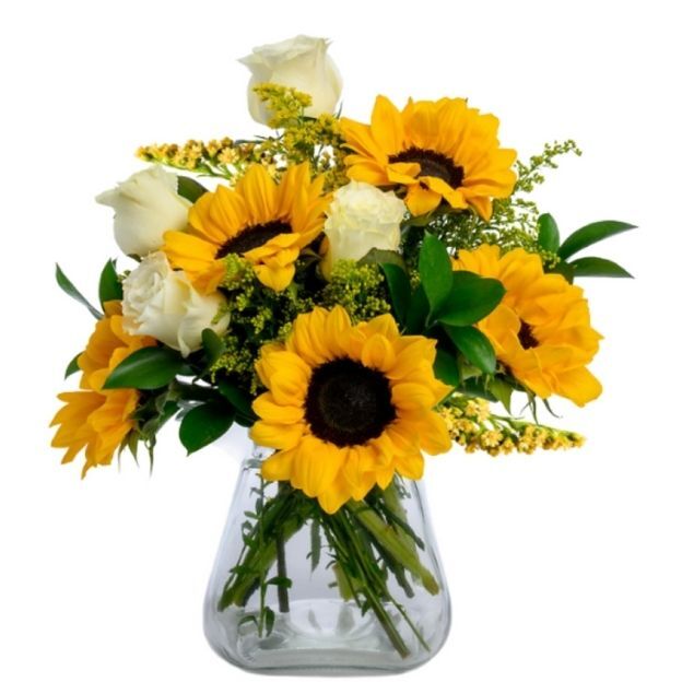 Bright sunflower and ivory rose bouquet with lush greens, stems visible in a wide clear vase.