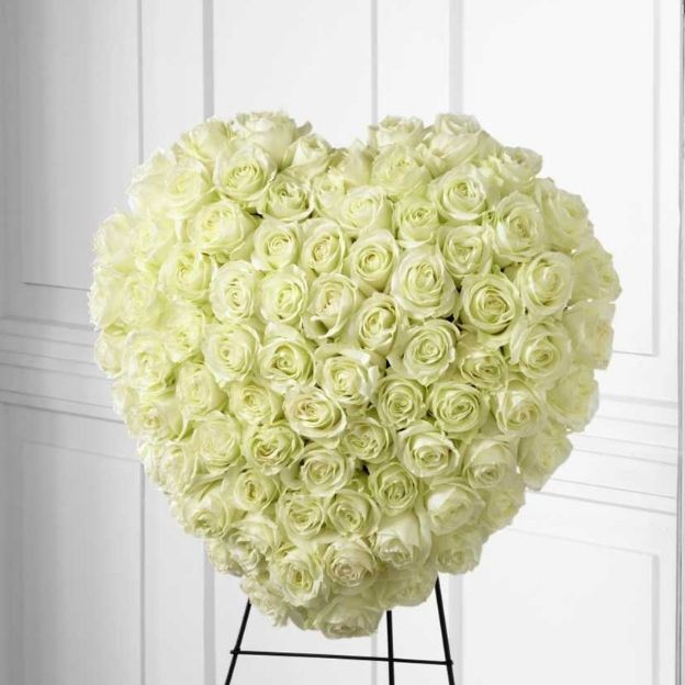 Heart-shaped standing spray of white roses on an easel, dense blooms forming a memorial wreath.