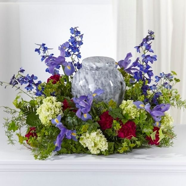 Memorial urn centerpiece with blue-purple delphinium, irises, red roses, and green hydrangea arranged in a ring.