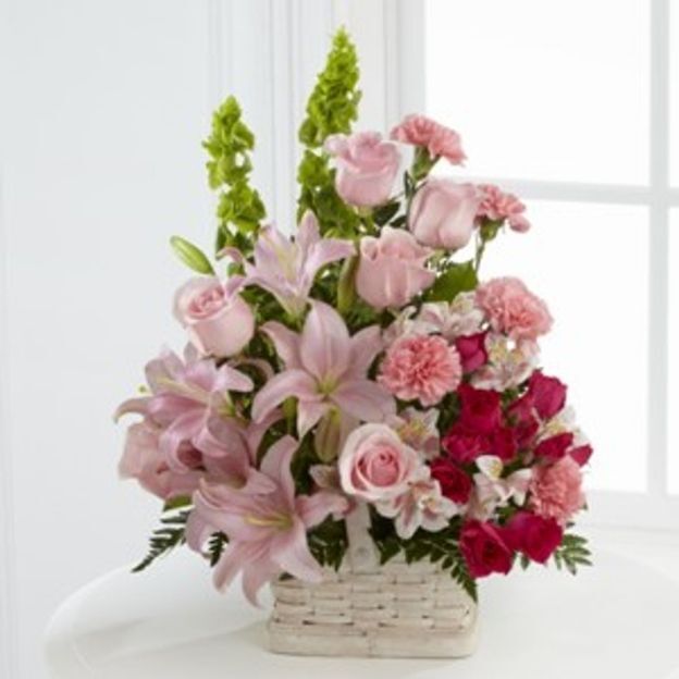 Soft pink lilies, roses, and carnations with green bells of Ireland, arranged in a white wicker basket.