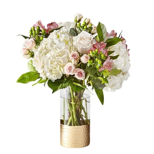 Elegant floral arrangement with white and blush flowers, green berries, and leaves in a tall vase with gold accent.