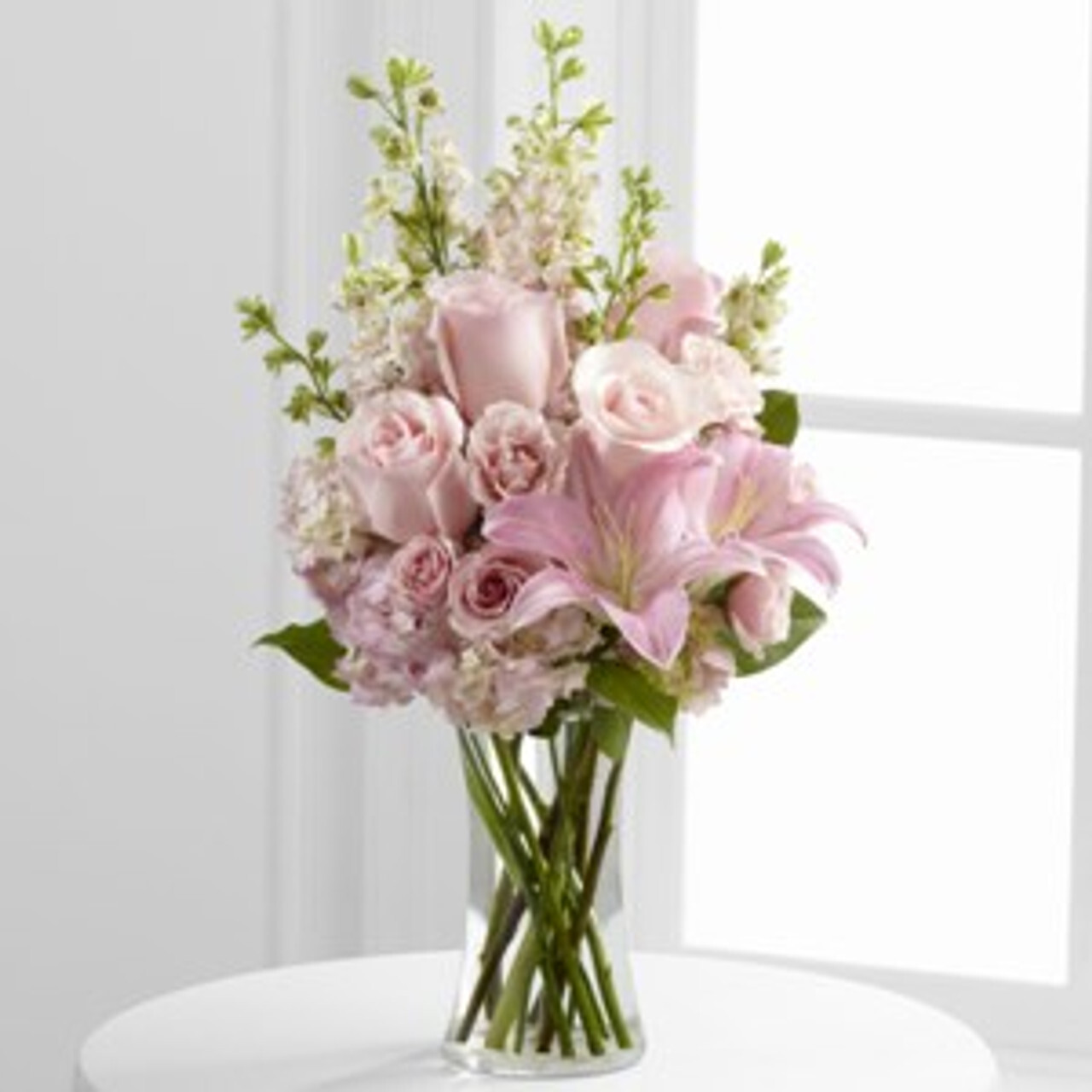 Elegant bouquet of pink roses, lilies, and stock flowers in a clear glass vase on a white table by a window.